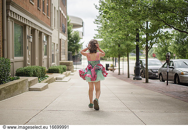 A small girl walks down sidewalk in tutu holding hair against wind