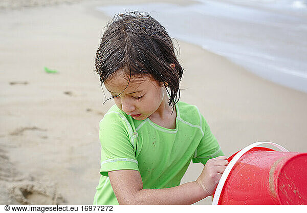 A small girl with wet sandy hair plays with a bucket on sandy beach
