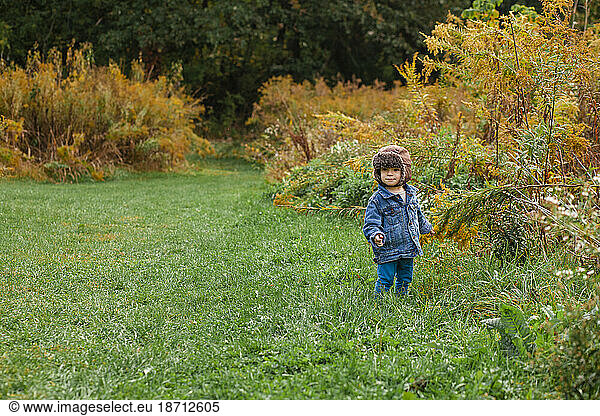 A small happy toddler stands bundled up on a grassy path in autumn