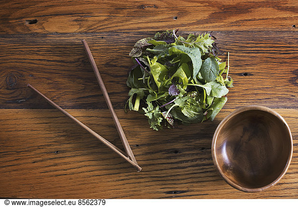 A small round polished wooden bowl and a clutch of organic mixed salad leaves  with wooden chopsticks.