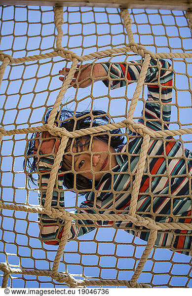 A smiling child looks drown through a rope net against a blue sky