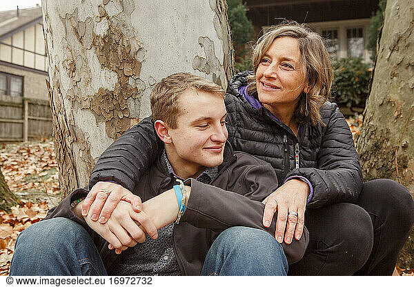 a smiling man sits against tree and wraps arm around teenage son