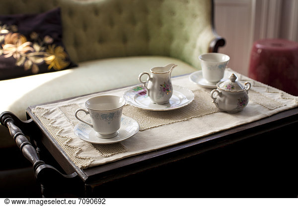 A sofa in a living room  and a tea tray. Mismatched cups and saucers  a china sugar bowl with a lid. Traditional design.