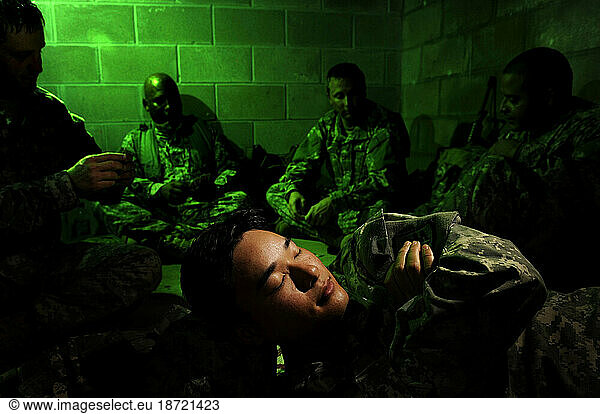 A soldier sleeps in a dark basement with very little light. Other soldiers relax and play cards in the background.
