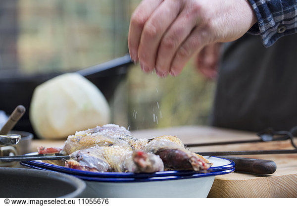 A spatchcocked game bird being seasoned for cooking  a man's hand sprinkling salt on the skin.