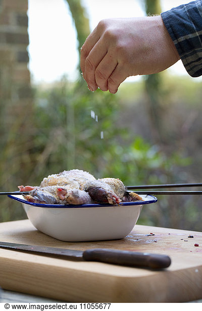 A spatchcocked game bird being seasoned for cooking  a man's hand sprinkling salt on the skin.