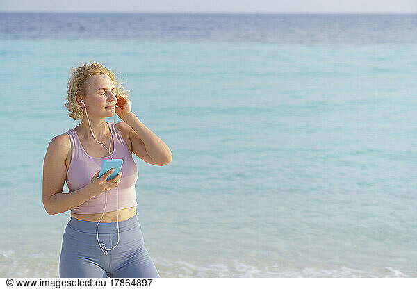 A sporty woman on the beach uses a smartphone and headphones.
