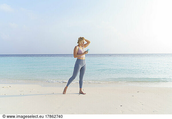 A sporty woman on the beach wearing headphones uses a smartphone.