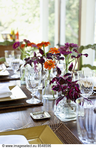 A table laid for a meal with glasses and glass vases of vivid coloured orange and maroon flower as a centrepiece.