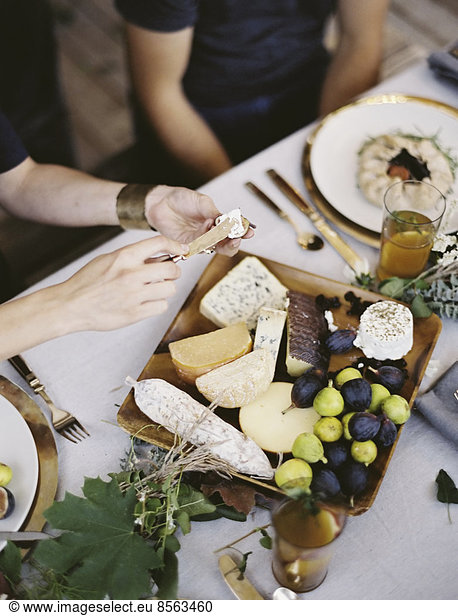 A table laid with a white cloth and place settings seen from above. An organic cheese board with soft and hard cheeses and figs. Two people sitting at the table.