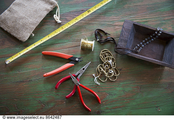 A tabletop with jewellery making equipment. Pliers  thread and a wooden tray  with measuring tape.