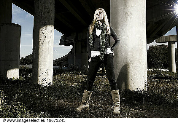 A tall blond female wears tall boots  black slacks and a scarf under and overpass.
