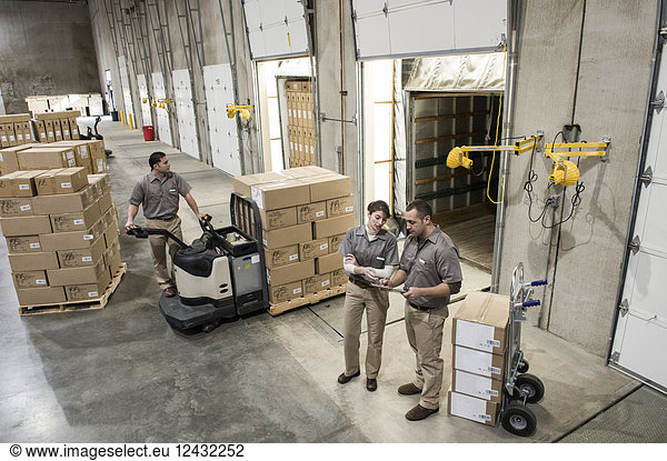A team of three mixed race uniformed warehouse workers loading boxed products into the back of a truck in a distribution warehouse.