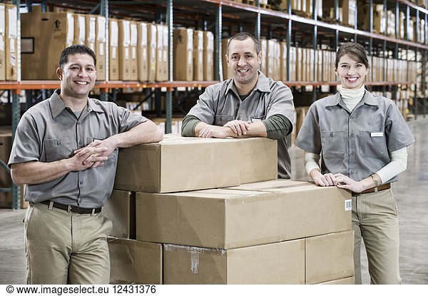 A team portrait of three mixed race uniformed warehouse workers surrounded by boxed products in a large distribution warehouse.
