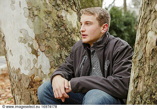 A teenage boy sits outside leaning against a sycamore tree in autumn