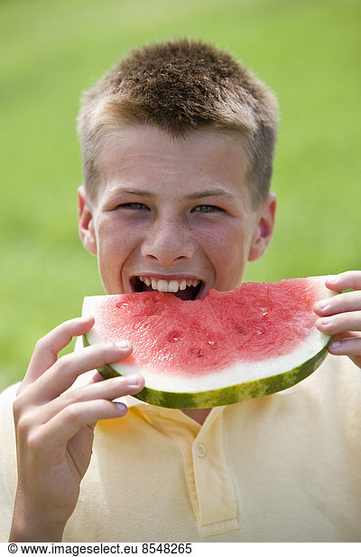 A teenage boy taking a large bite out of a watermelon.