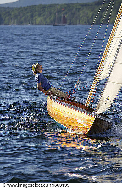 A teenaged boy happily sailing a classic wooden sailing dinghy  looks up to check the sails  on a glorious summer day.