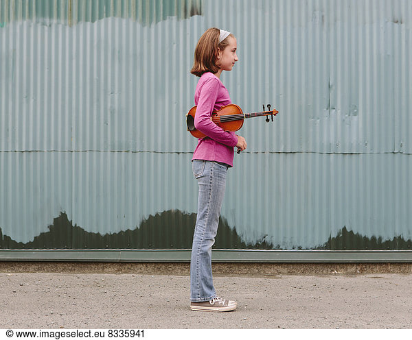 A ten year old girl holding a violin under her arm  standing on a street.