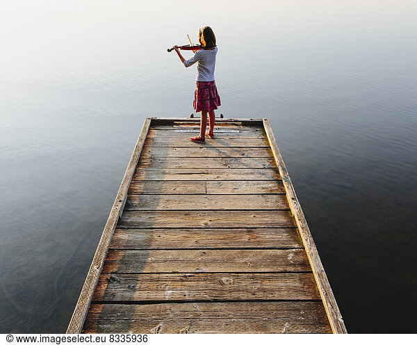 A ten year old girl playing the violin at dawn on a wooden dock.