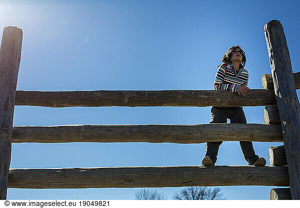 a thoughtful child stands on a high wooden fence against blue sky