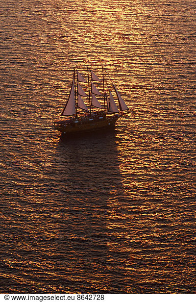 A three-masted sailing ship with full sail on the Aegean Sea at sunset.