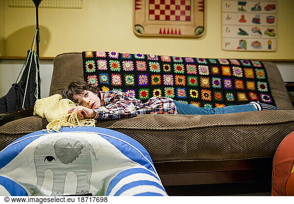 A tired child rests on a living room couch with a multicolored blanket