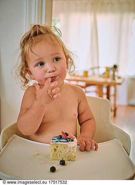 A toddler licks her fingers while eating birthday cake.