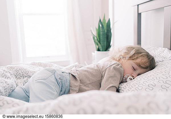 A toddler sleeping in her parents bed with a pacifier.