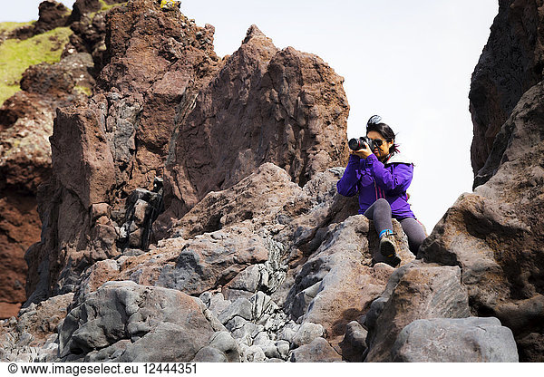 A tourist sits on the top of a rock formation to take a higher photo of the landscape  Iceland
