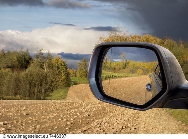 A view of the road behind a vehicle in a car's side mirror  A rural landscape with forests and farmland