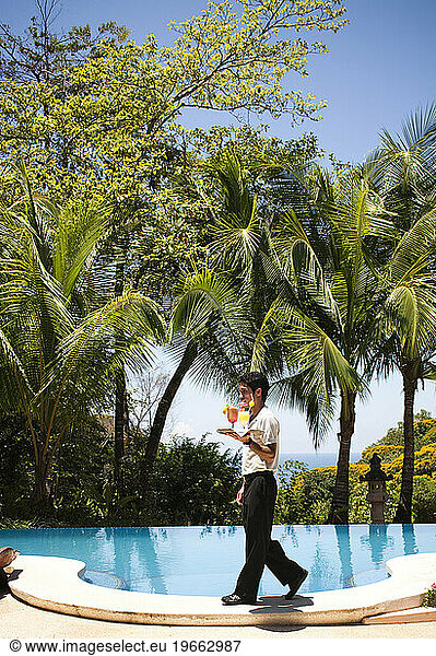 A waiter walks past an outdoor pool on a sunny day to deliver two drinks.
