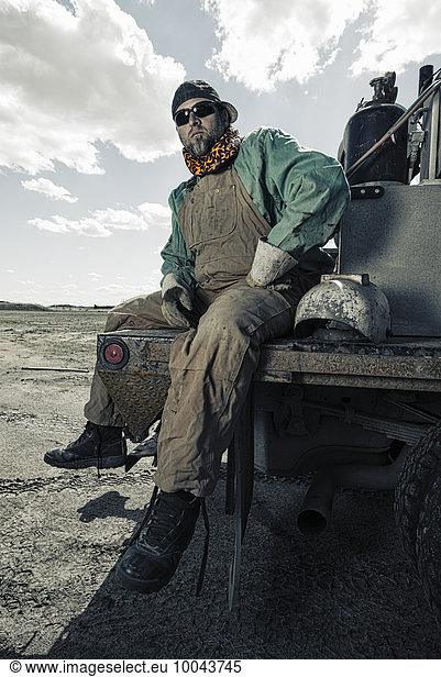 A welder seated on a truck with his gear beside him.