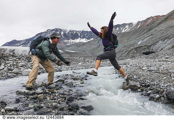 A wife jumps to her husband across an icy stream while hiking near Gulkana Glacier  Alaska  United States of America