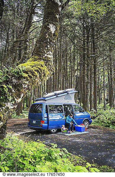 A woman and small child sit outside camper van in forest