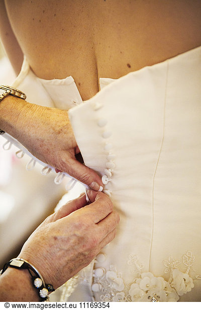 A woman buttoning the back of a bride's white wedding dress.