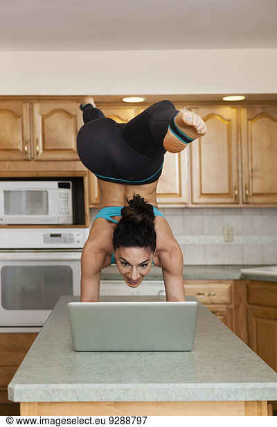 A woman doing a handstand balancing on a kitchen top and checking her laptop.