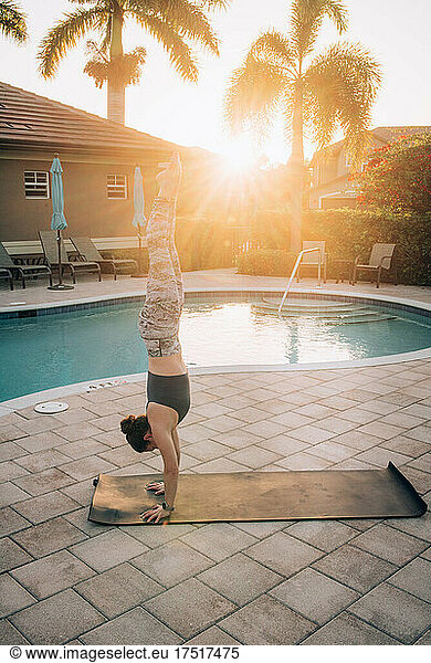 A woman doing a handstand & mat pilates next to a pool at sunrise