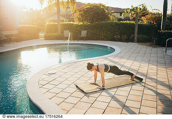 A woman doing mat pilates & planks next to a pool at sunrise
