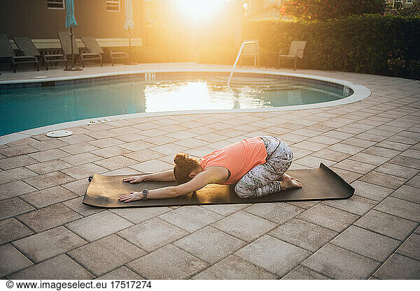 A woman doing mat pilates & stretching next to a pool at sunrise