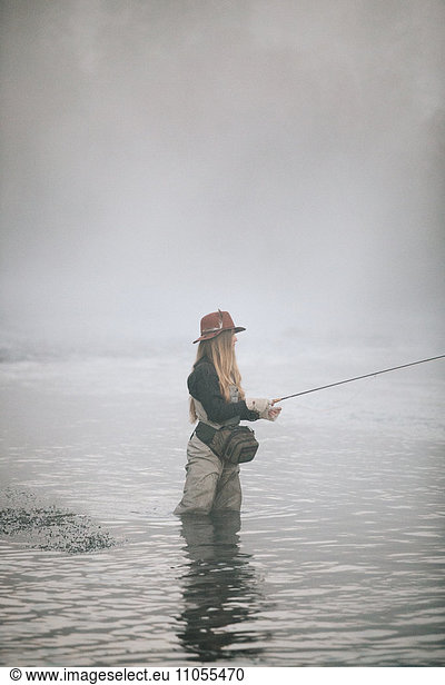 A woman fisherman fly fishing  standing in waders in thigh deep water.