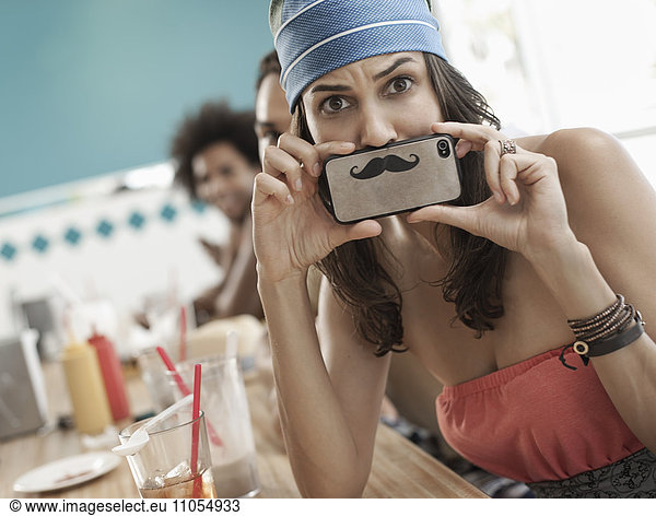 A woman holding a picture of a moustache on her smart phone just under her nose.
