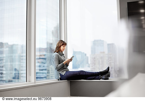 A woman holding a smart phone  sitting on a window ledge.
