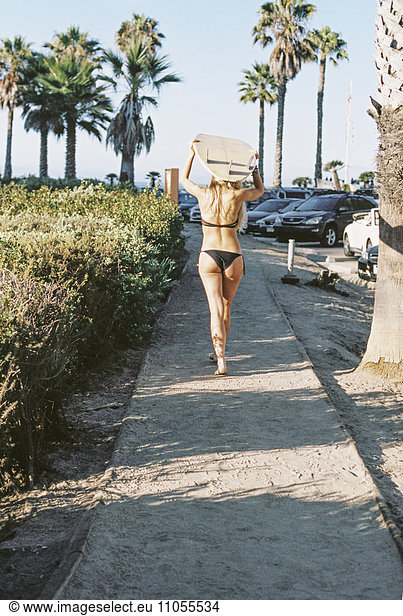 A woman in a black bikini walking down a path carrying a surfboard on her head.