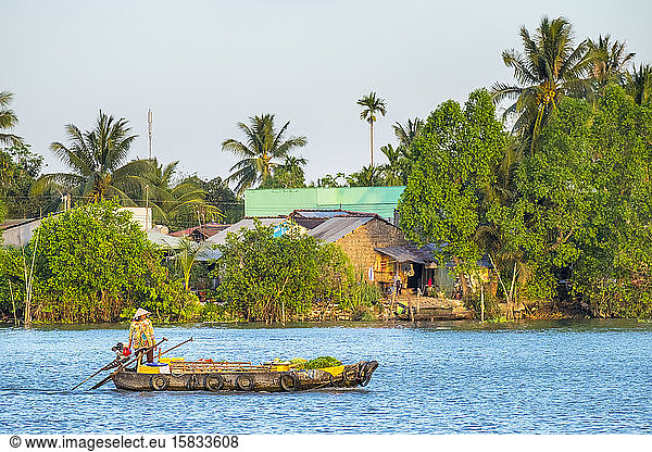 A woman in a boat passes a village on a branch of the Mekong River