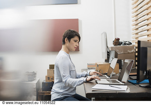 A woman in a home office seated at a desk with two laptops  her hands on the keyboard of one computer  looking at the screen on the other.