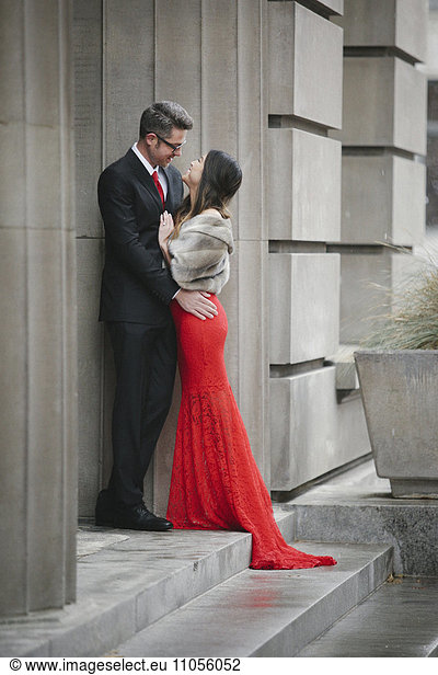 A woman in a long red evening dress with fishtail skirt and a fur stole  and a man in a suit  embracing on the steps of a building.