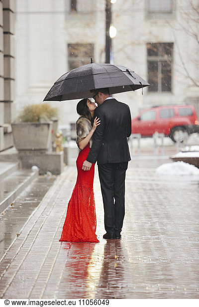 A woman in a long red evening dress with fishtail skirt and a fur stole  and a man in a suit kissing under an umbrella on a street.