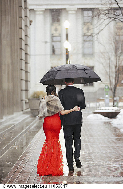 A woman in a long red evening dress with fishtail skirt and a fur stole  and a man in a suit  walking through a city.