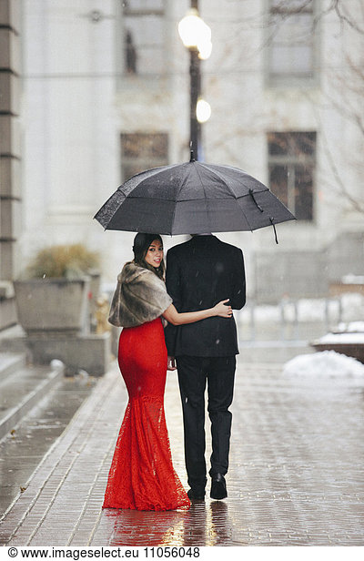 A woman in a long red evening dress with fishtail skirt and a fur stole  and a man in a suit  walking through snow in the city.