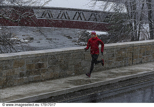 A woman in a red jacket running in winter near a covered bridge.